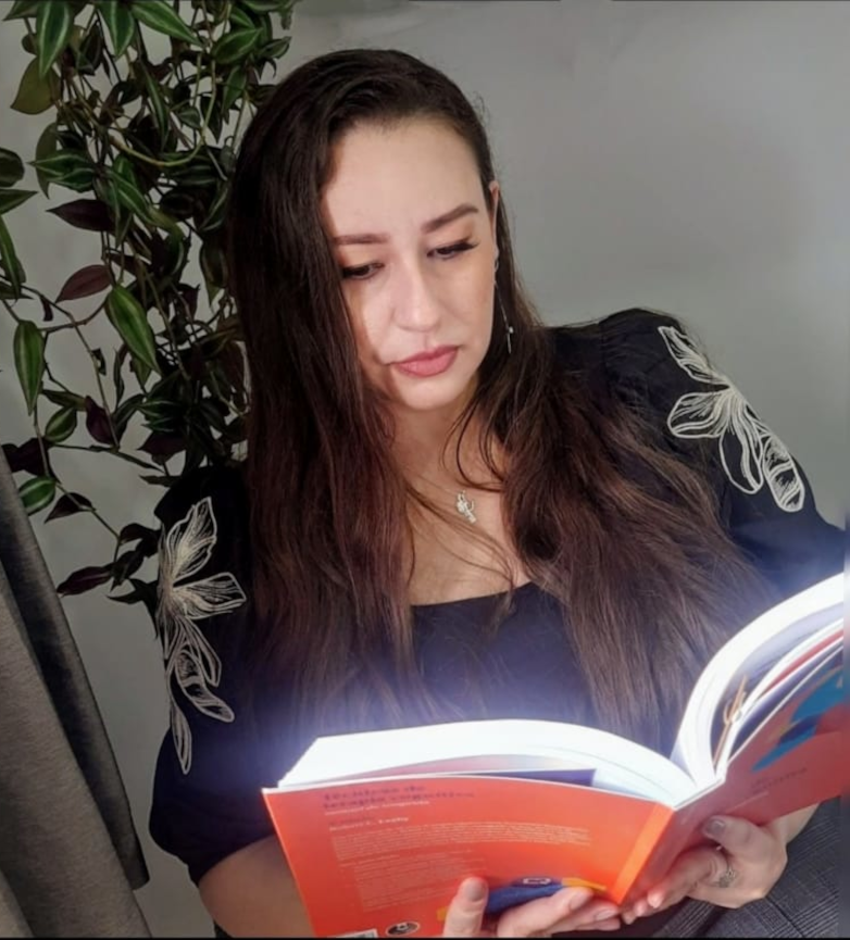 Psicóloga Fernanda com expressão séria, lendo um livro com planta ao fundo